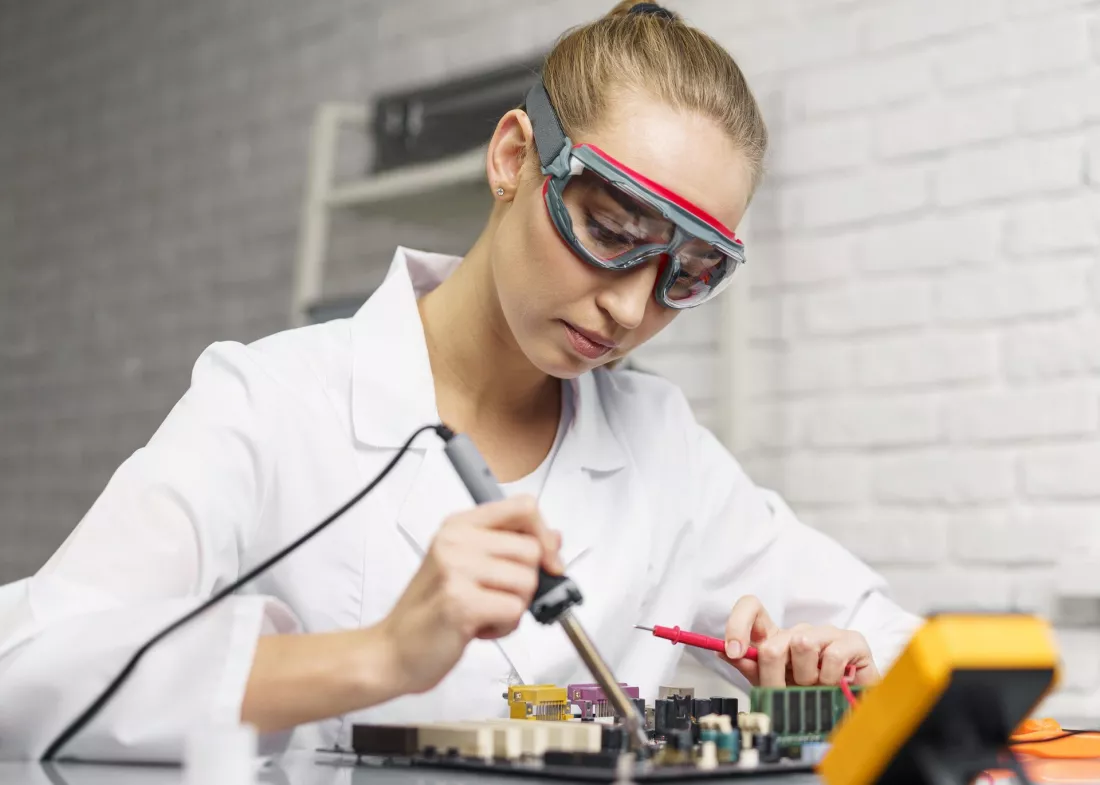 Studentka w okularach ochronnych lutuje elementy na płytce elektronicznej, obok leży multimetr